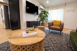 A modern living room with a round wooden coffee table, a sofa with a yellow cushion, a television mounted on the wall, and a large potted plant.