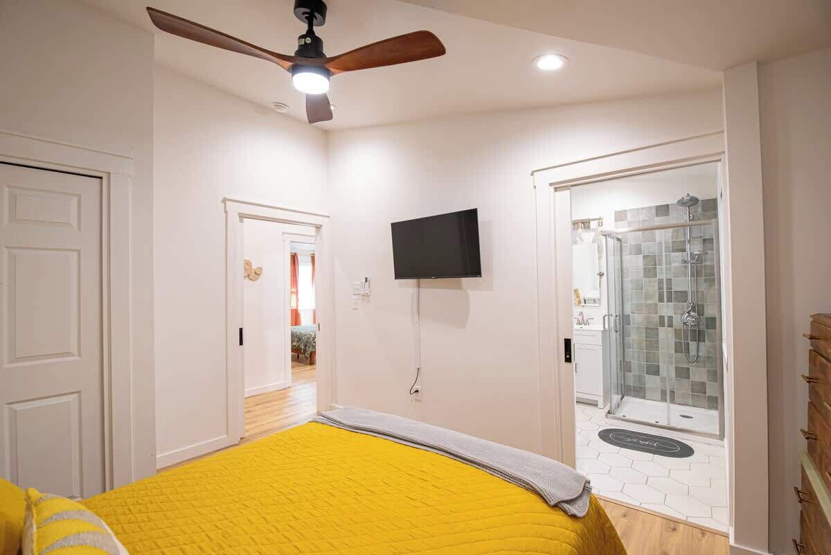 A bedroom with a bed, a ceiling fan, a TV, and a shower area.