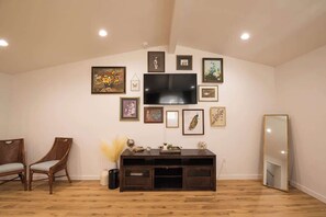 Ein Zimmer mit an der Wand befestigtem Fernseher, umgeben von gerahmten Bildern, einer Holzkommode und zwei Korbstühlen.