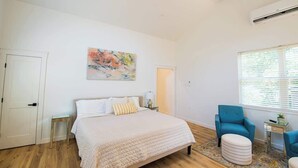 Un dormitorio con cama, una silla, una mesita, un cuadro y una ventana con persianas.