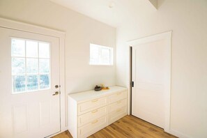 Quarto com porta branca, janela, um banco branco e chão de madeira.