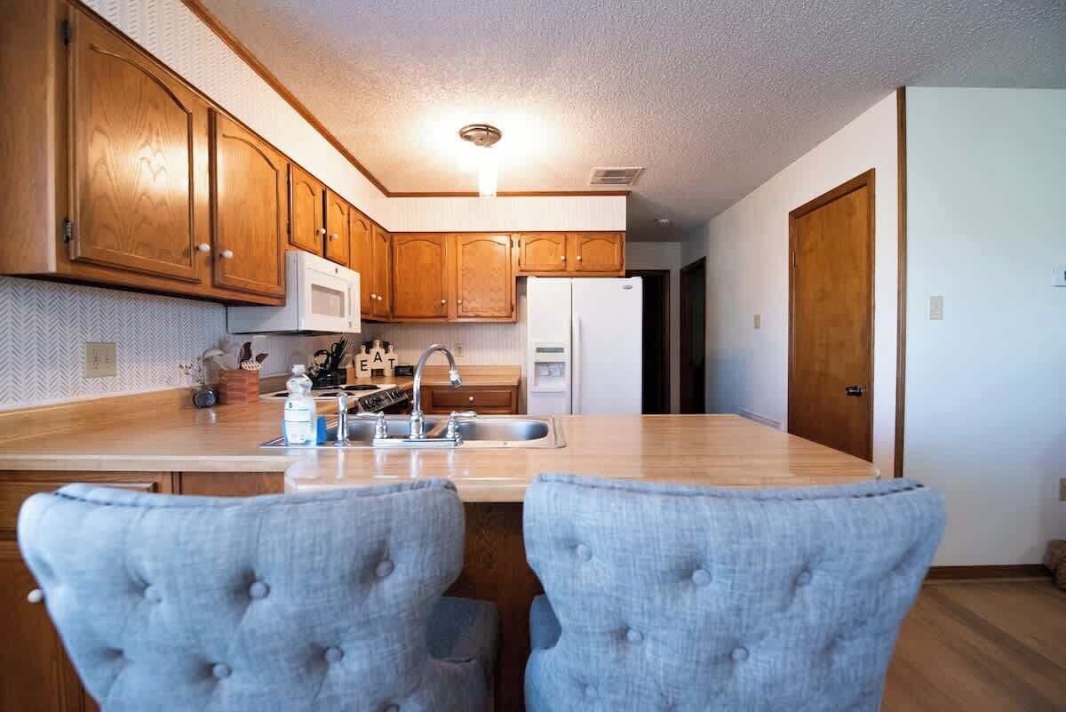 A kitchen with wooden cabinets, a microwave, a refrigerator, a sink, and two upholstered chairs.