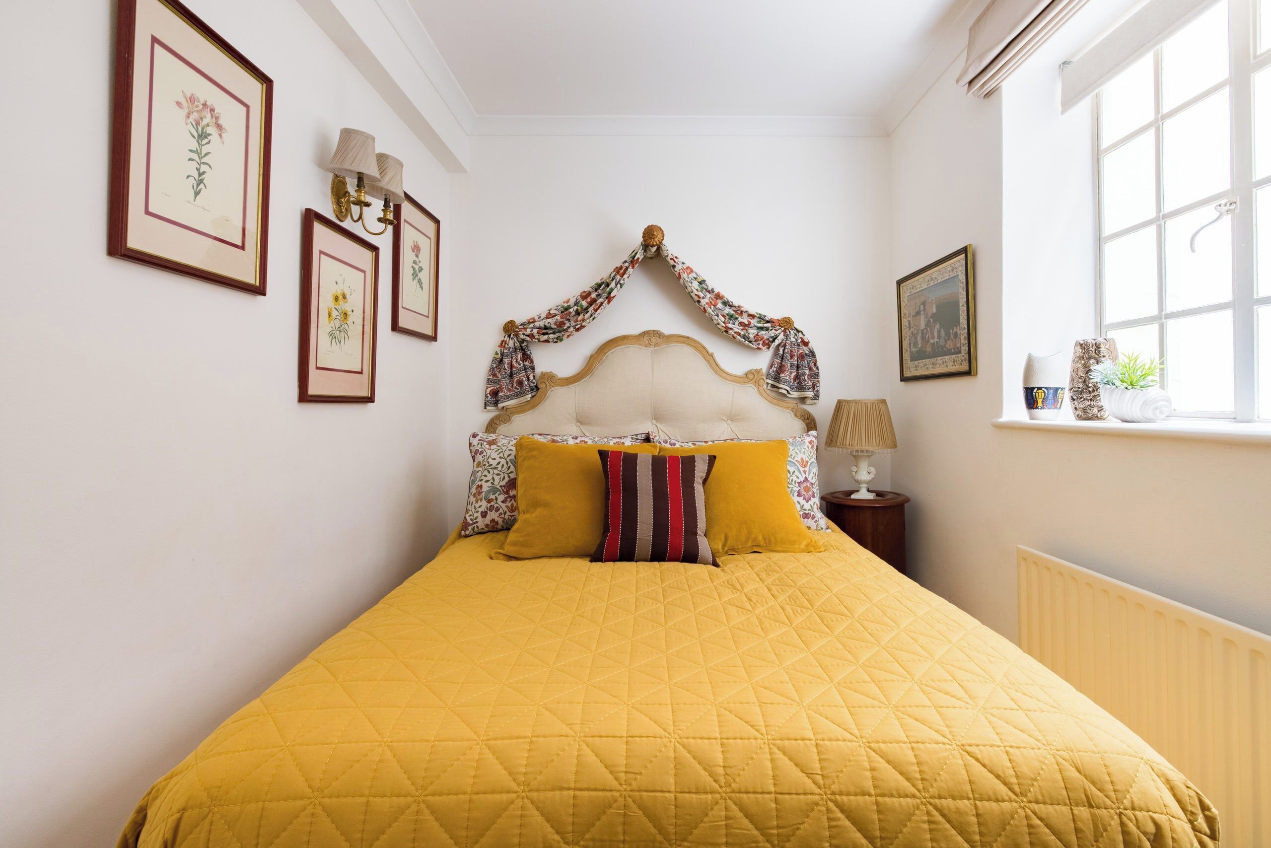 A bed with a yellow quilt, a headboard with a floral curtain, and framed floral artwork on the wall.