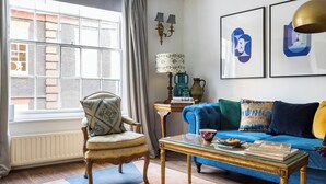A living room with a blue sofa, a patterned armchair, a glass coffee table, and a window with a view of a brick building.