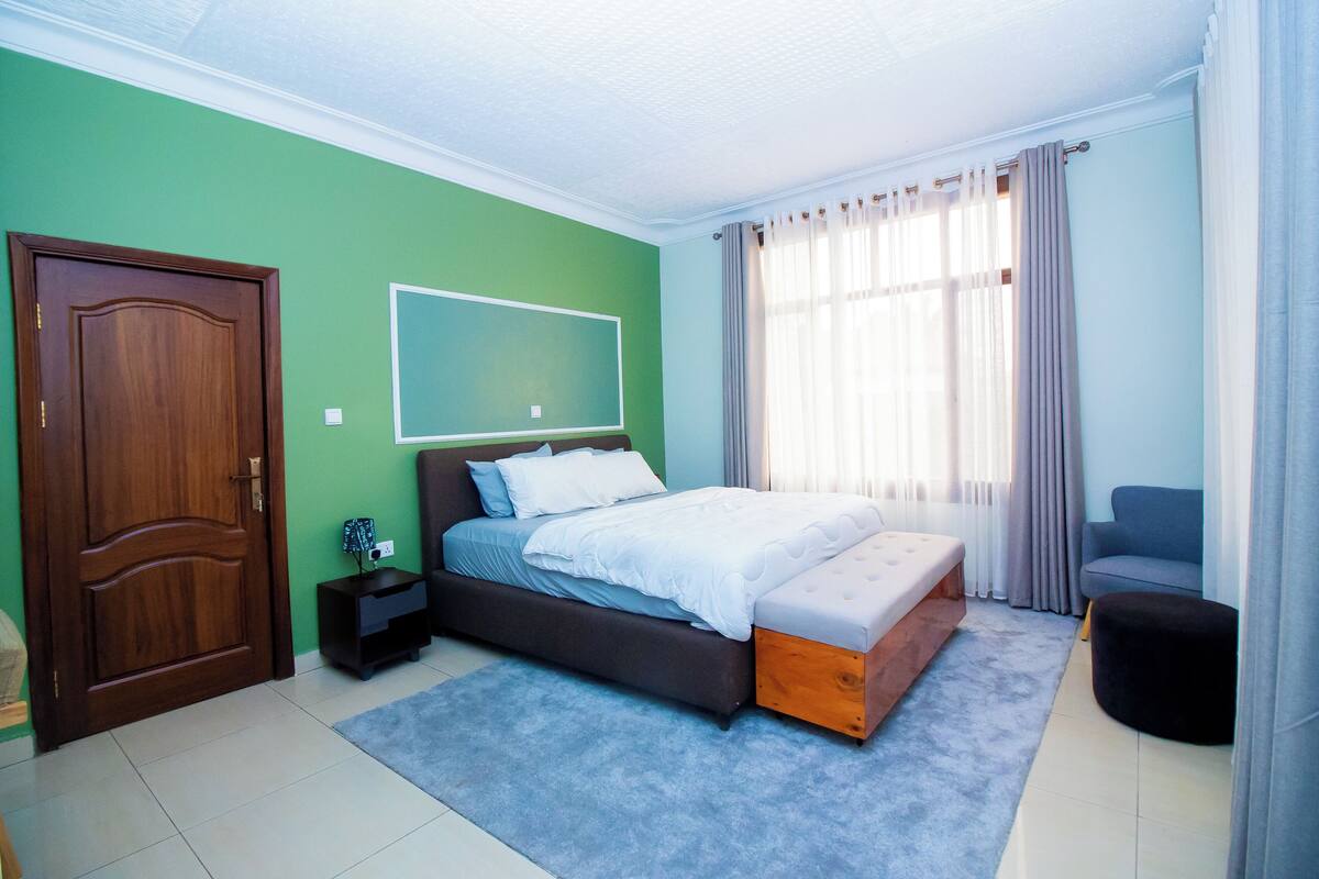 A bedroom with a bed, a nightstand, a chair, a window with curtains, and a door.