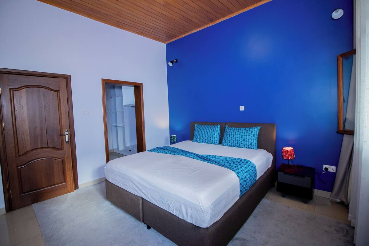 A bedroom with a large bed, a wooden door, a mirror, and a nightstand with a lamp.