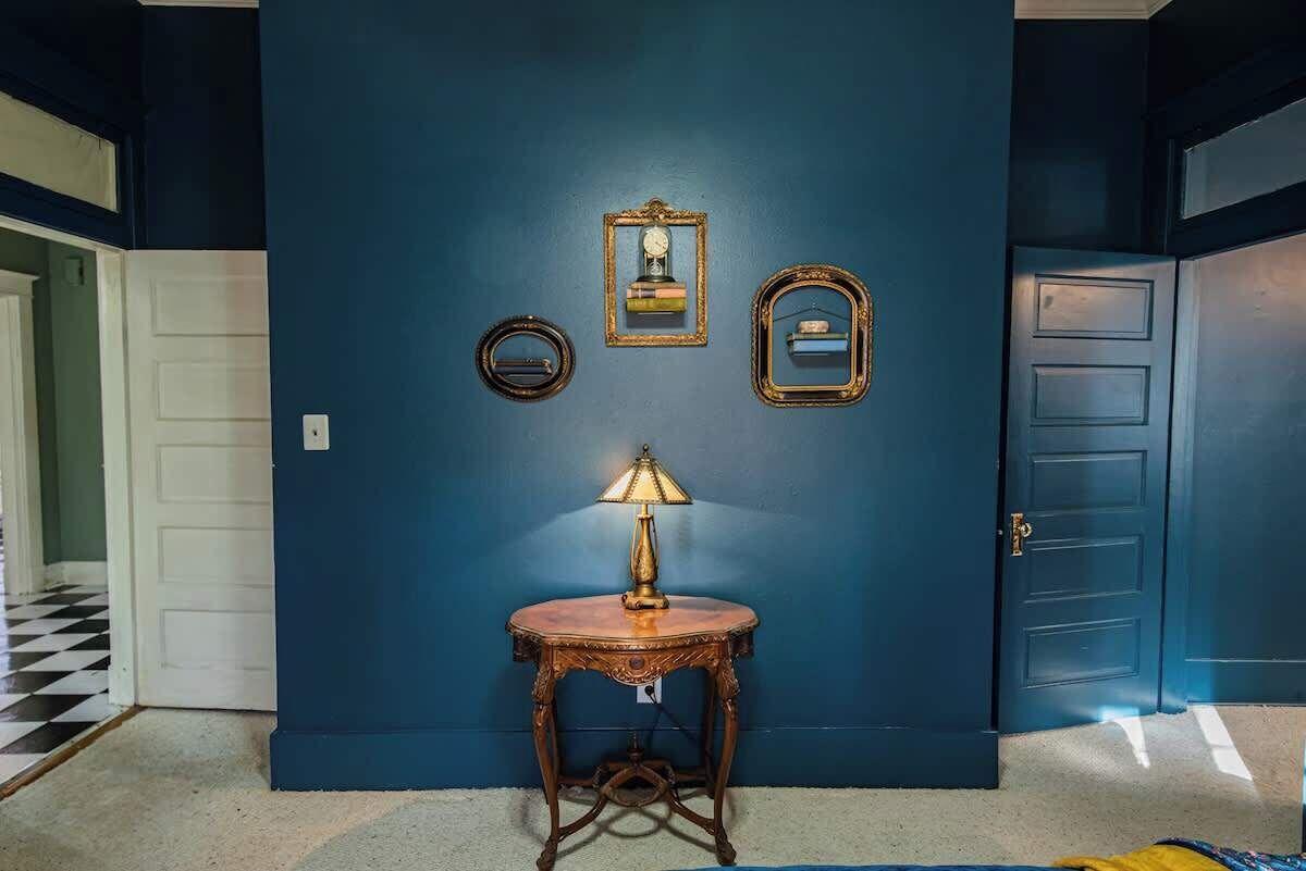 A room with a blue wall, a round wooden table with a lamp, and three framed items above the table.