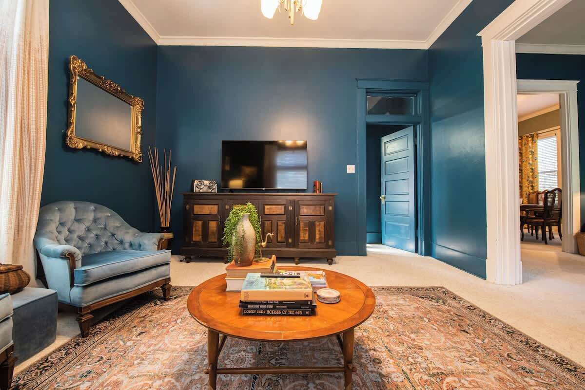 Ein Wohnzimmer mit blauer Wand, einem Fernseher, einem blauen Sofa, einem runden Couchtisch und einem gemusterten Teppich.