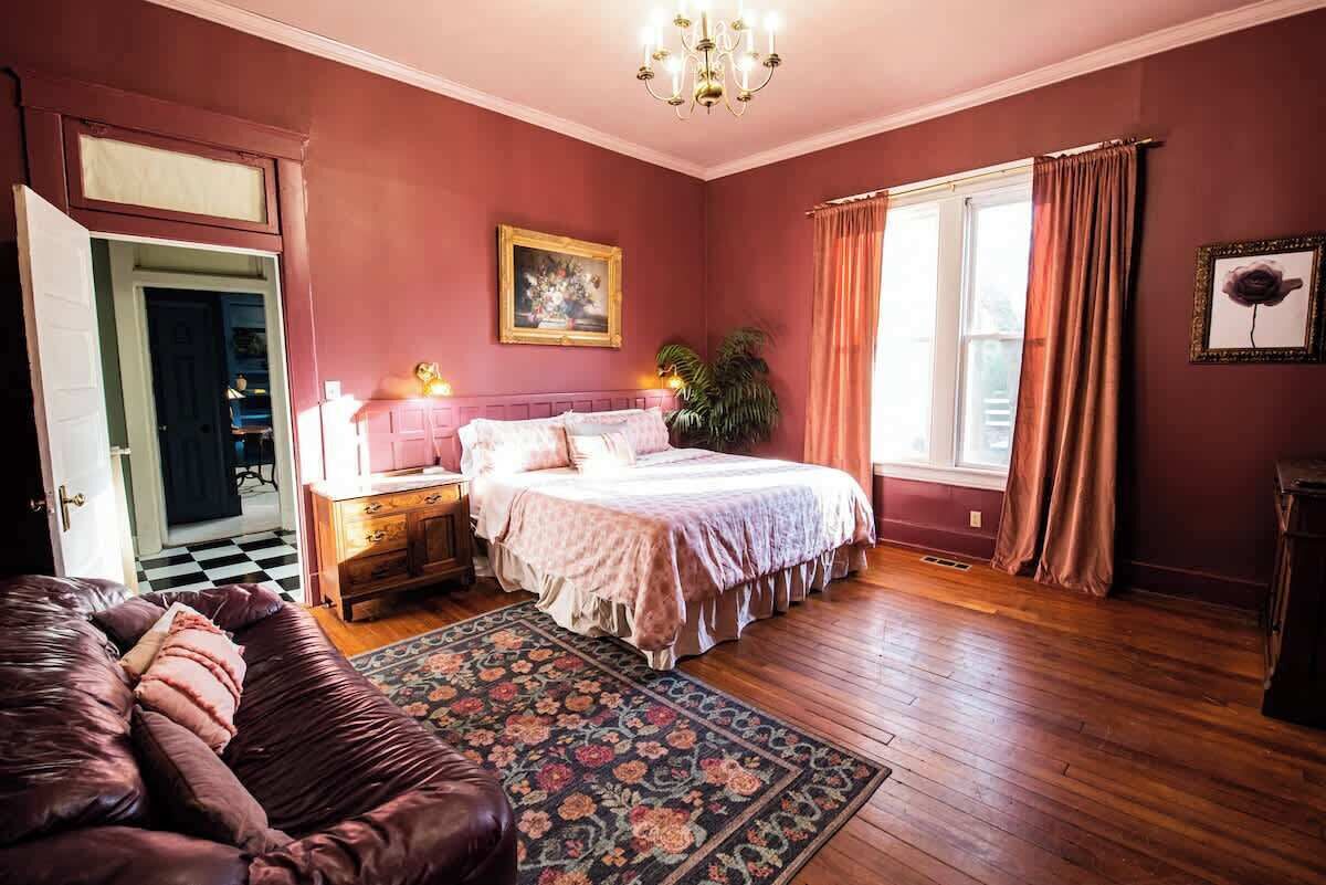 A bedroom with a bed, nightstands, a dresser, a chair, a window with curtains, and a chandelier.