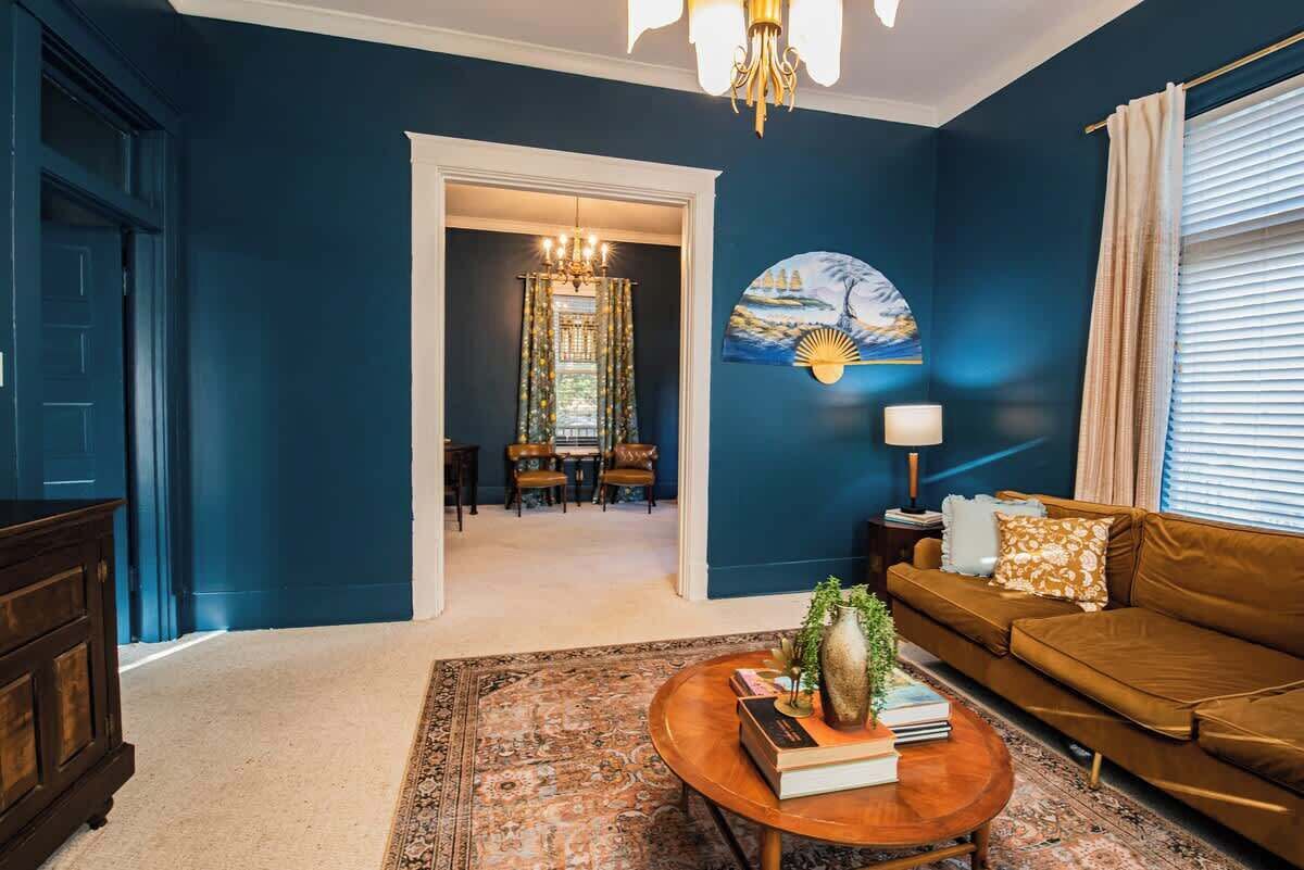 A room with a blue wall, a brown sofa, a coffee table with books and a vase, a chandelier, and a painting.