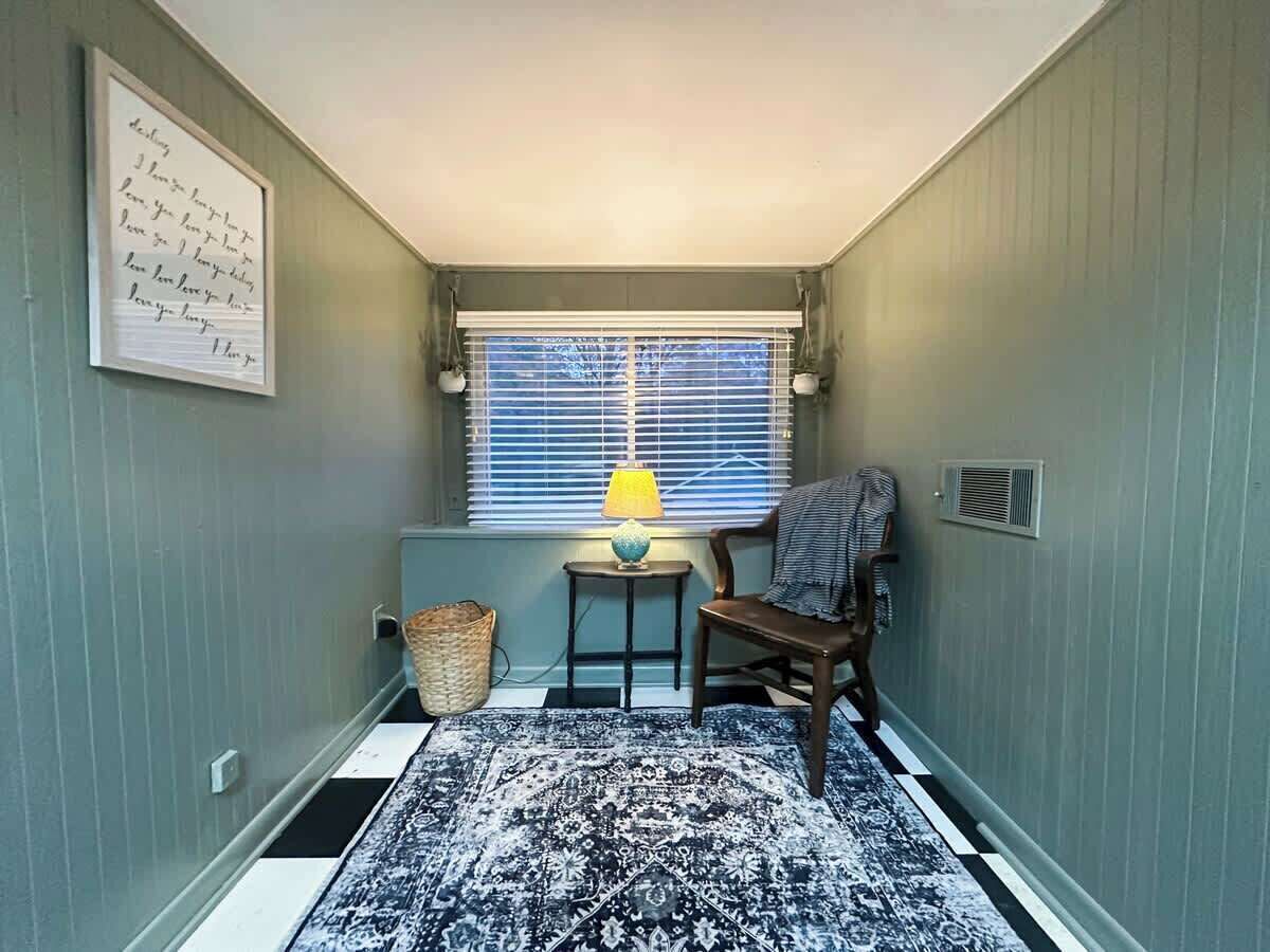 Une petite pièce avec une fenêtre, une lampe, une chaise, un tapis et un interrupteur fixé au mur.