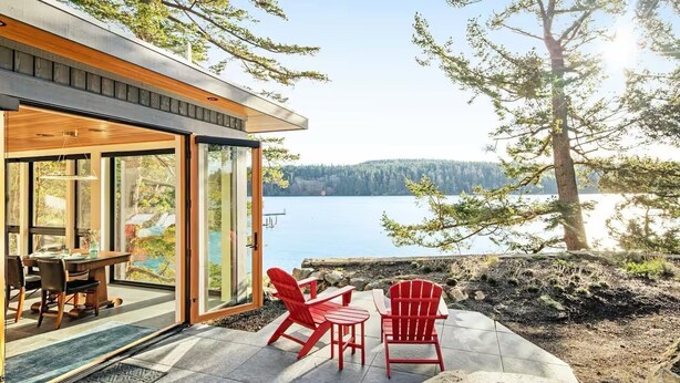 Une maison moderne avec une porte vitrée donnant sur un patio équipé d’une table et de deux chaises rouges, le tout offrant une vue sur un lac.