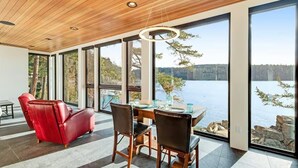 Une pièce spacieuse avec un plafond en bois, de grandes fenêtres donnant sur un lac, une table à manger avec des chaises et un canapé en cuir rouge.