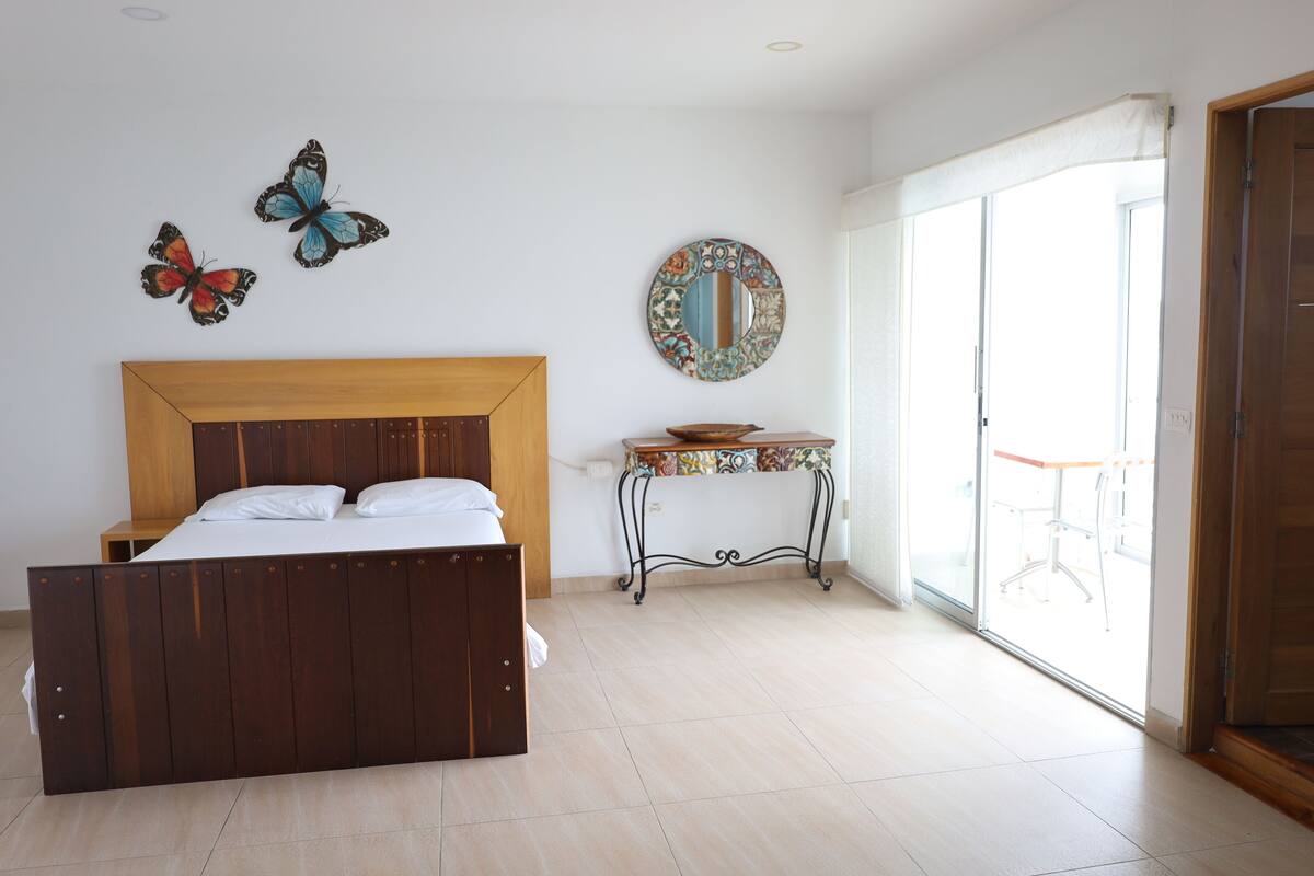 Ein Schlafzimmer mit einem hölzernen Bett, einem kleinen Tisch mit einer bunten Ablage, einem dekorativen Spiegel und Wandbildern von Schmetterlingen.