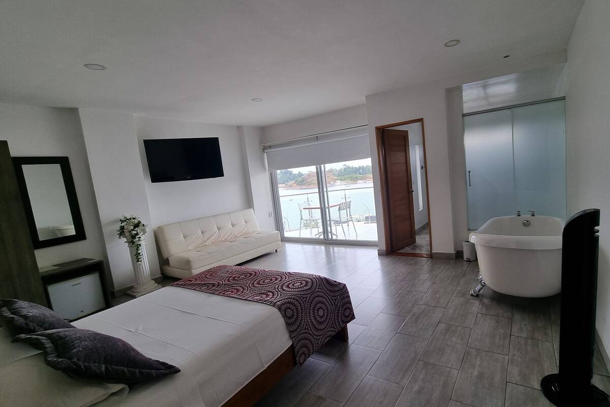 Ein Schlafzimmer mit Bett, Fernseher und einem Badezimmer mit Badewanne, mit Blick auf das Meer durch eine Glastür.