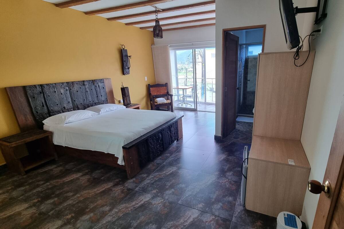 Ein Schlafzimmer mit einem hölzernen Bett, einem Nachttisch, einem Kleiderschrank und Blick ins Freie durch eine Glastür.