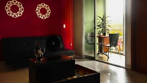 A living room with a red wall, a black sofa, a coffee table with a bottle and glasses, and a sliding glass door leading to a balcony with a plant and a chair. - HOTEL PORTOBELO GUATAPE (Guatapé)