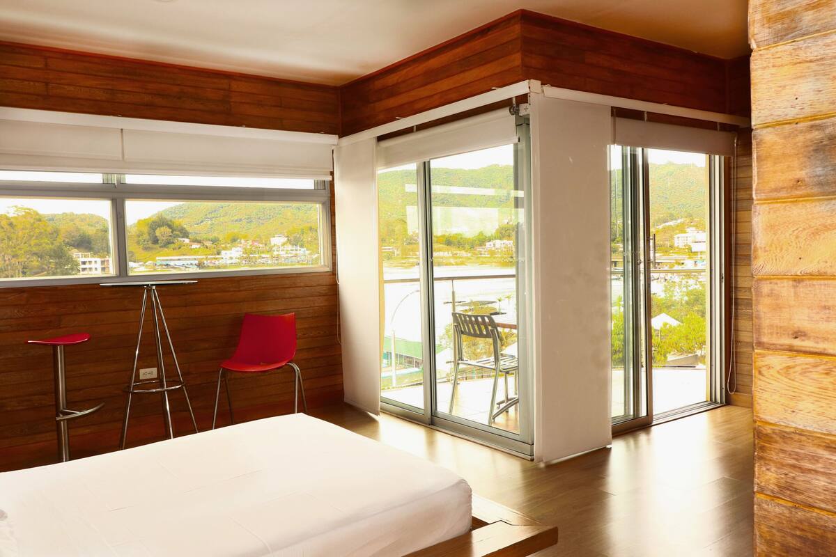 Ein modernes Hotelzimmer mit einem Bett, einem Essbereich mit roten Stühlen und einer Glastür zu einem Balkon mit Blick auf Grünflächen und Gebäude.