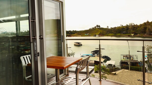 A balcony with a wooden table and chairs, overlooking a body of water with boats and a hilly landscape. - HOTEL PORTOBELO GUATAPE (Guatapé)