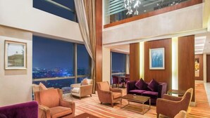 Eine moderne Hotelhalle mit hoher Decke, großen Fenstern mit Blick auf die Stadt und verschiedenen Sitzgelegenheiten, darunter Sofas und Sessel.