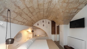 A bedroom with a stone ceiling, a bed with white bedding, a bedside lamp, a wall-mounted TV, and a small shelf with decorative items. - Palazzo Fiscaiolo (Monopoli)