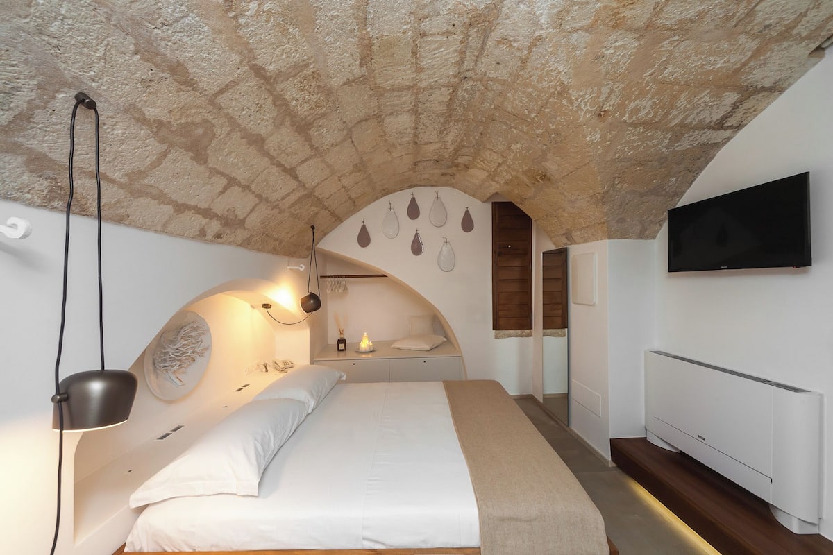 A bedroom with a stone ceiling, a bed with white bedding, a bedside lamp, a wall-mounted TV, and a small shelf with decorative items.