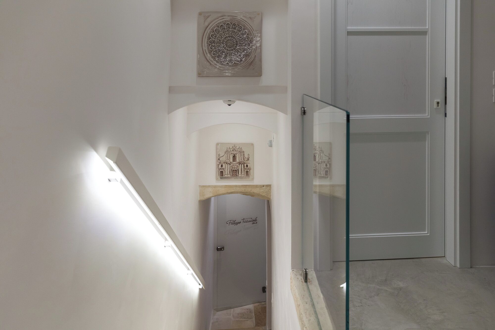 A hallway with a glass door, a white wall with a decorative plate, and a staircase leading to a door with a plaque.