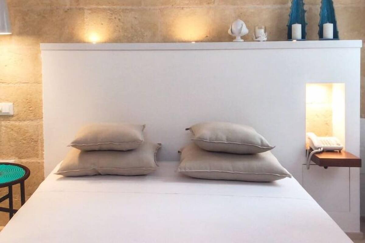 A bed with two pillows, a small bedside table with a lamp, and a wall-mounted shelf with decorative items.