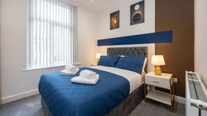 A neatly made bed with blue bedding and white towels, a bedside table with a lamp, and a radiator. - CollegeHouse-FREEParking&HighSpeedWIFI-Modern-Cosy (Whalley Range)