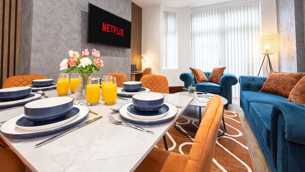 A modern hotel room with a dining area, a blue sofa, and a flat-screen TV displaying 'NETFLIX'. - CollegeHouse-FREEParking&HighSpeedWIFI-Modern-Cosy (Whalley Range)