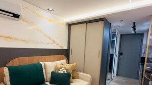 Un salon moderne avec un canapé, une table basse et un climatiseur fixé au mur. On y trouve une porte donnant sur une autre pièce et un couloir avec un miroir et une porte.
