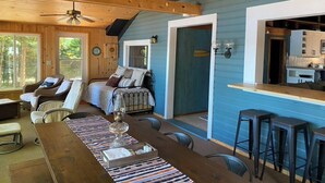 A cozy cabin interior with a dining area, a seating area with a sofa and chairs, and a kitchen visible through an open doorway. - Adorable Lakefront Cabin with HOT TUB! (Gresham)