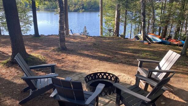 A campsite with two Adirondack chairs facing a fire pit, surrounded by trees and a lake. - Adorable Lakefront Cabin with HOT TUB! (Gresham)
