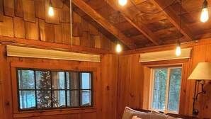 A cozy wooden cabin bedroom with a bed, a window with a view of trees, and a lamp. - Adorable Lakefront Cabin with HOT TUB! (Gresham)