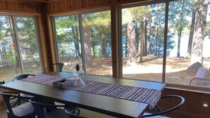 A wooden dining table with a placemat, surrounded by four chairs, in a room with large windows offering a view of a forested area. - Adorable Lakefront Cabin with HOT TUB! (Gresham)