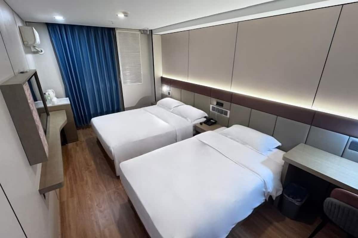 Habitación de hotel con dos camas, un escritorio, una silla, una mesita, un cubo de basura y una ventana con cortinas.