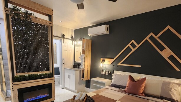 A modern bedroom with a large bed, a ceiling fan, a wall-mounted air conditioner, and a decorative wall panel. - Cozy Apartment at Citrus Park (Tampa)
