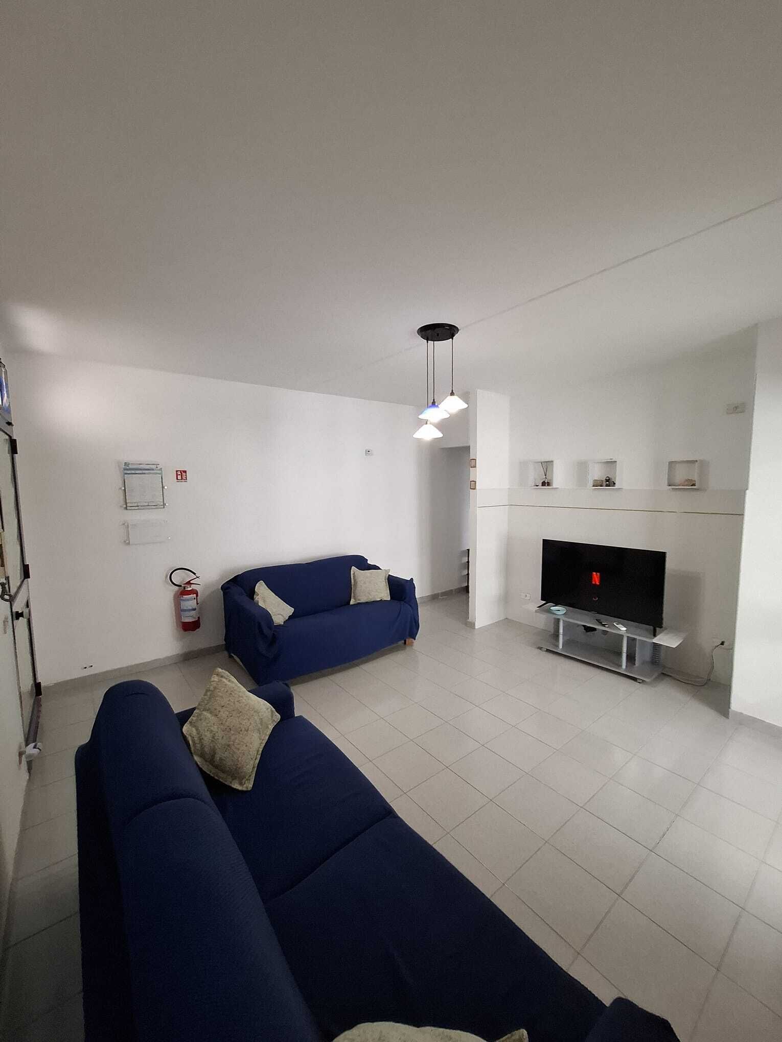 A modern living room with two blue sofas, a flat-screen TV, a ceiling light fixture, and a fire extinguisher on the wall.