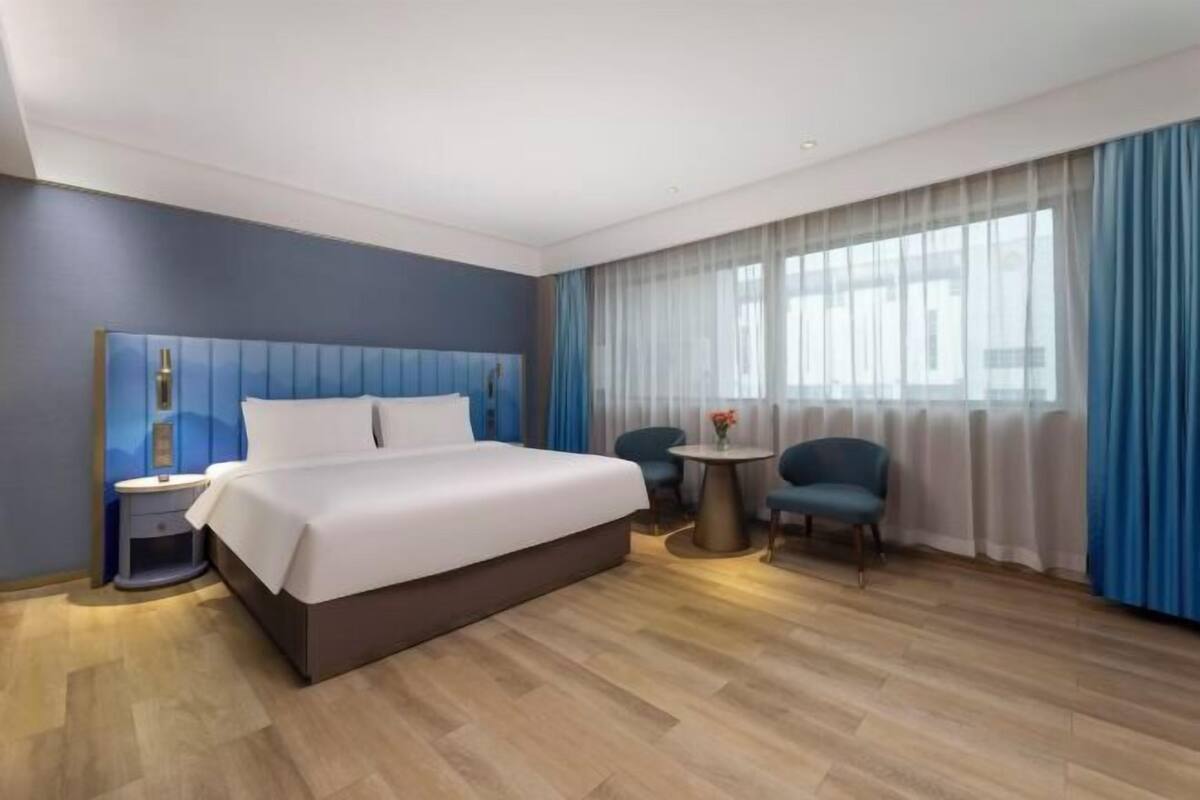 A modern hotel room with a large bed, a bedside table, a small round table, two chairs, and a large window with curtains.