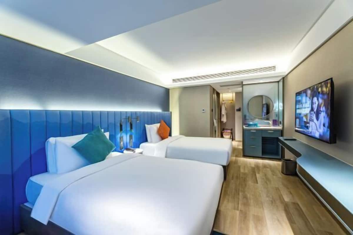 A modern hotel room with two beds, a TV, a mirror, and a small kitchenette.