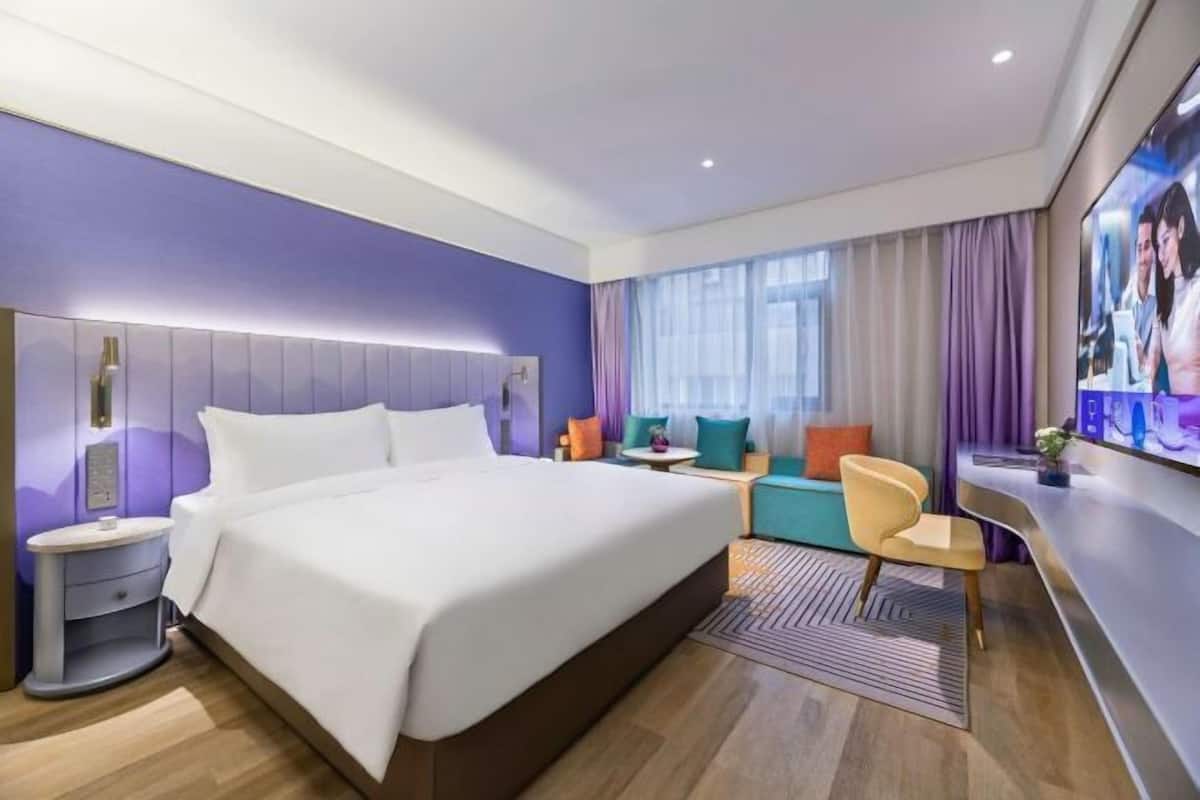 A modern hotel room with a large bed, a bedside table, a small desk, a chair, and a TV.