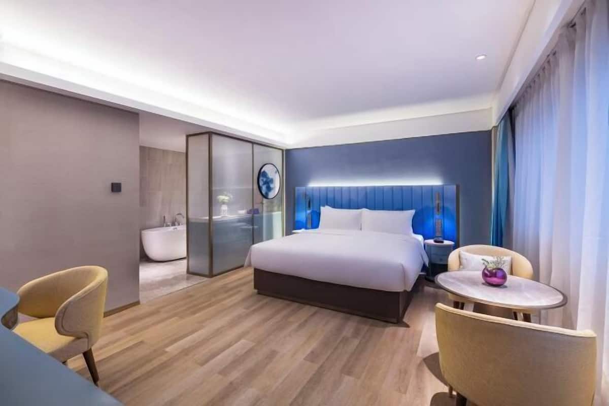 A modern hotel room with a large bed, a bedside table, a chair, and a small round table.