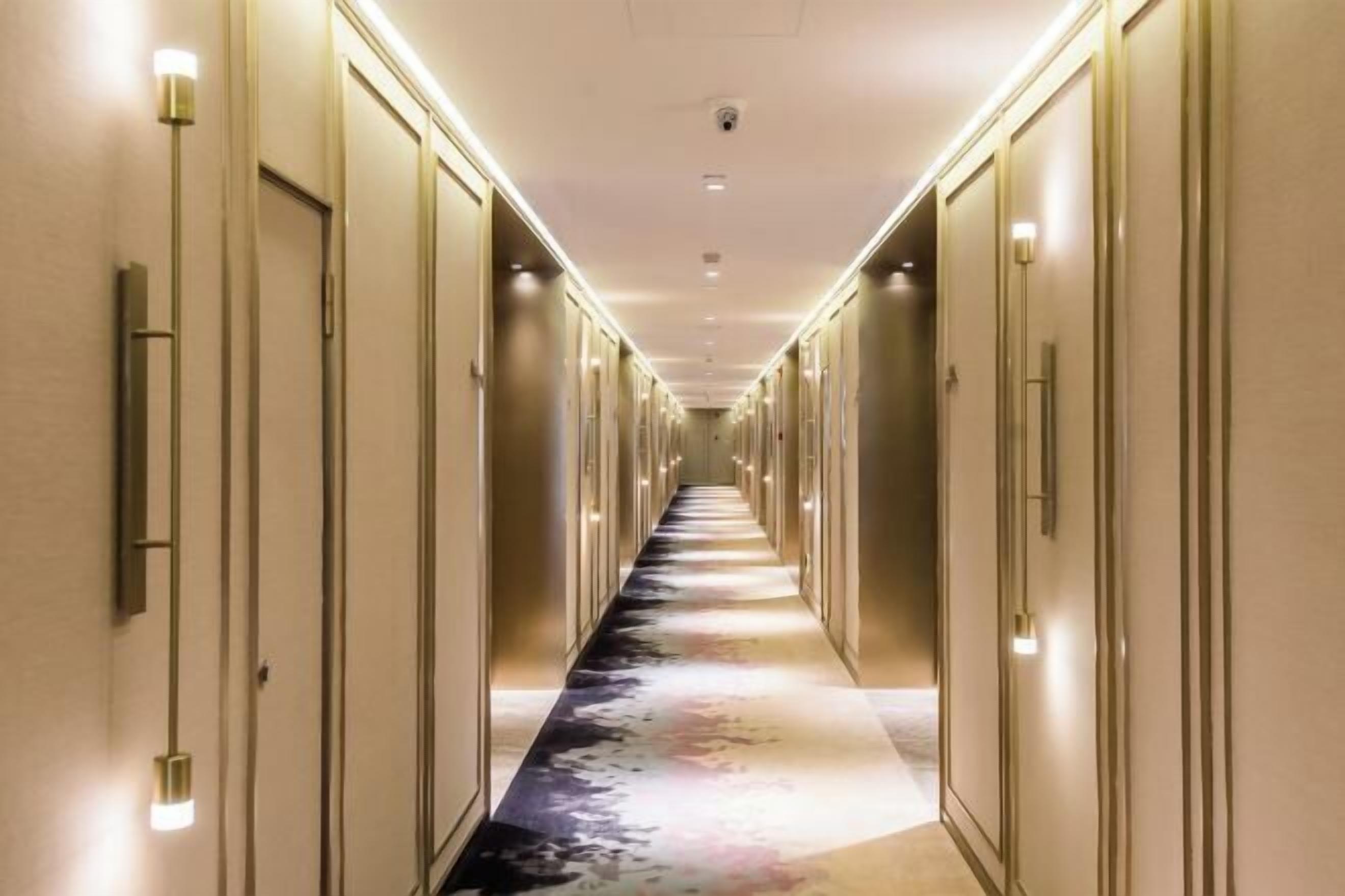 A long, narrow hotel corridor with doors on both sides, illuminated by overhead lights.