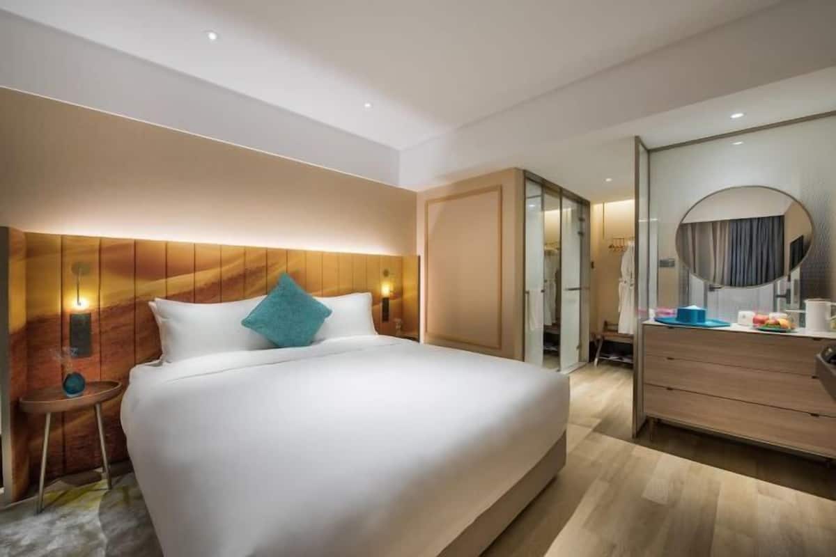 A modern hotel room with a large bed, a wooden headboard, a bedside table with a lamp, a dresser, and a mirror.