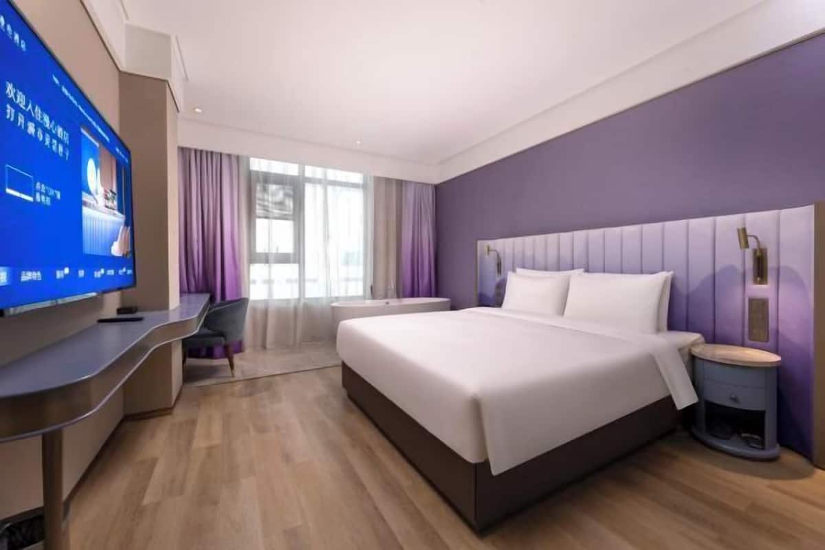A modern hotel room with a large bed, a desk, a television, and a window with curtains.