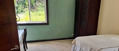 A bedroom with a bed, a wardrobe, a desk, and a window with a view of greenery.