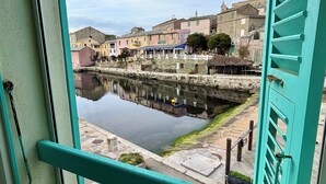 A view through a turquoise-framed window overlooking a calm water body with buildings in the background. - Casa Marelina - Fisherman's house in Centuri harbour (Centuri)