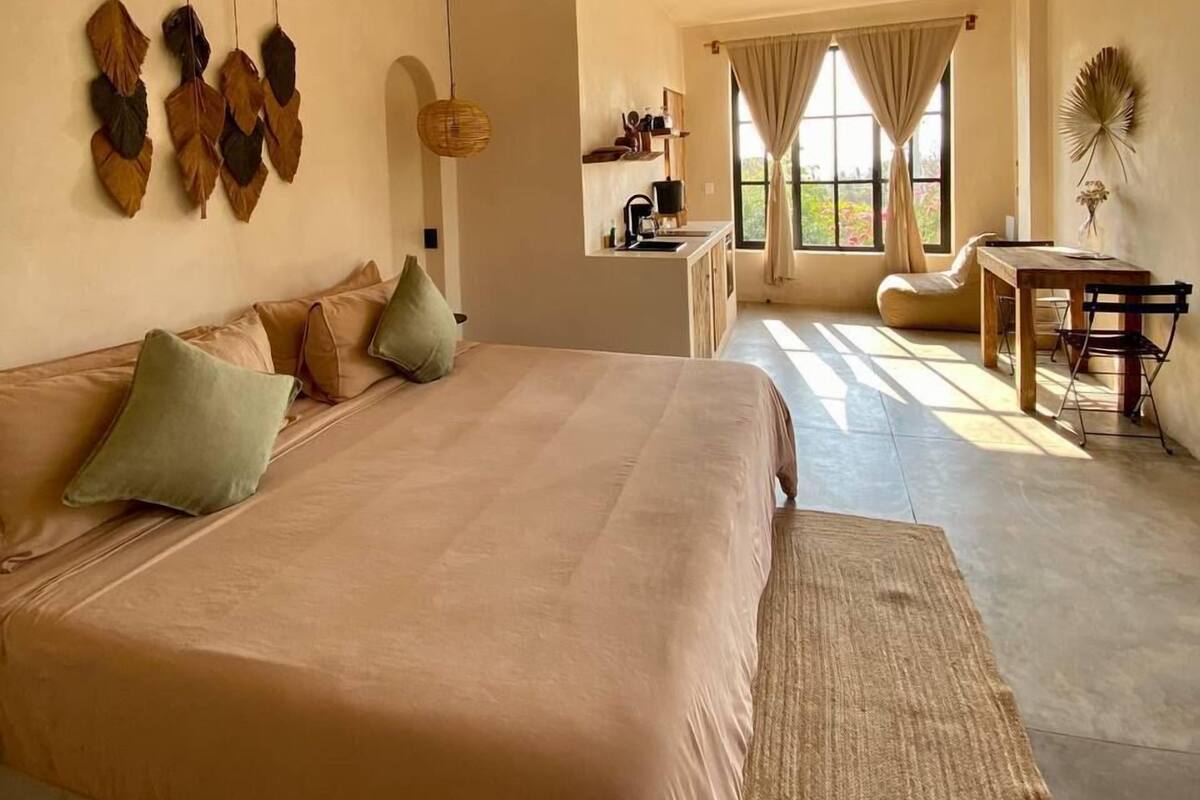 A spacious bedroom with a large bed, a ceiling fan, and a view of the kitchen and dining area through a window.