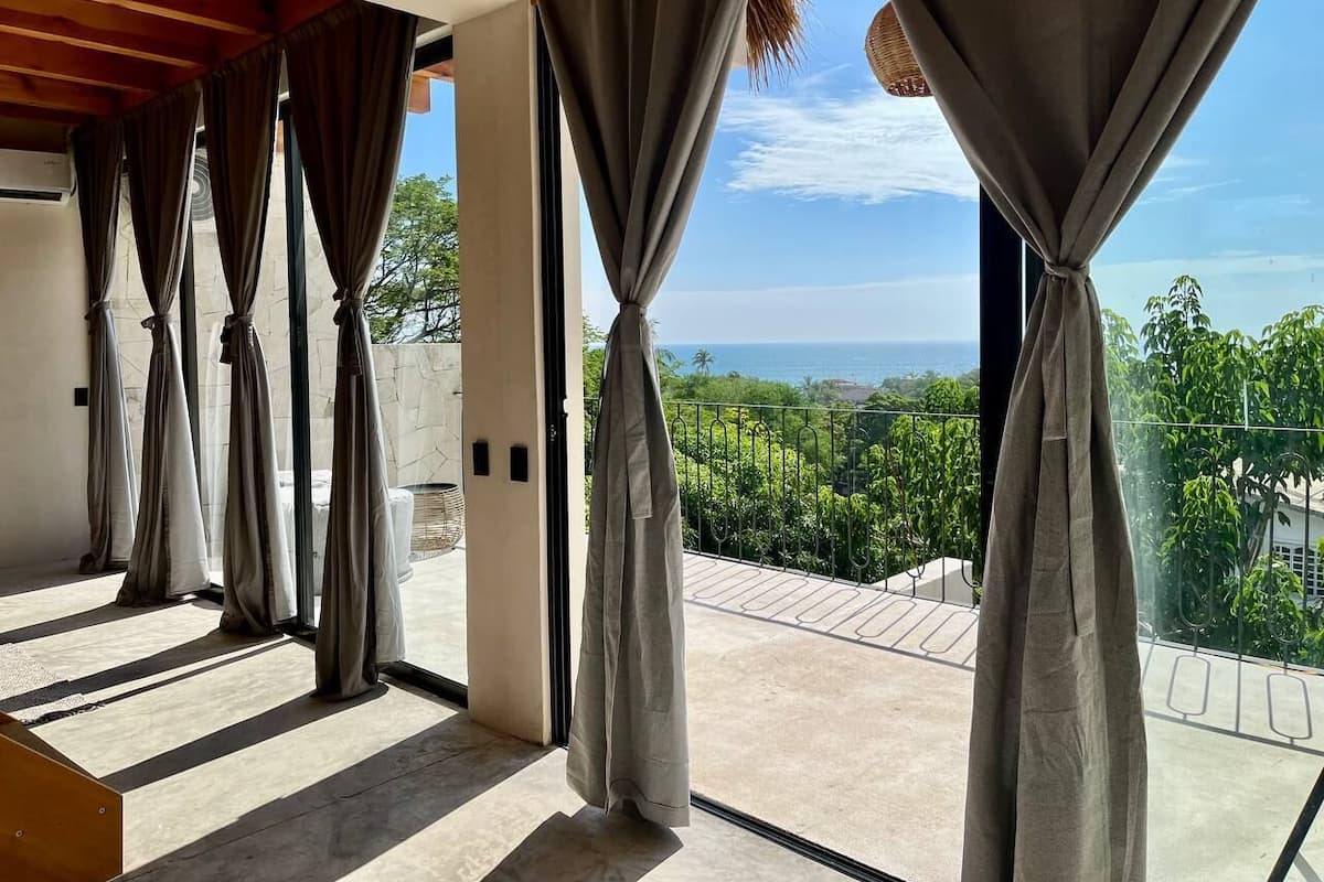 A room with large windows, a view of greenery and the sea, and a balcony with a metal railing.