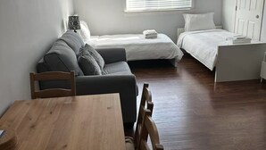 Een compacte kamer met een houten eettafel, twee bedden, een bank en een raam met zonwering.
